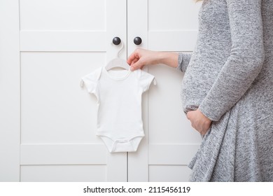 White Baby Bodysuit Hanging On Hanger At Door Of Wardrobe At Nursery Room. Young Adult Pregnant Woman With Big Belly Preparing Clothes For Future Newborn.  