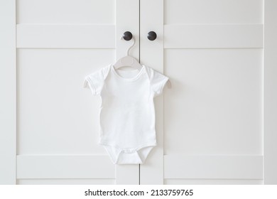 White baby bodysuit hanging on hanger at door of wardrobe. Closeup. Front view. Clothes preparing for newborn.