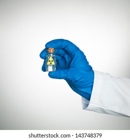 White Background With Hand In Blue Glove And White Lab Coat Holding A Small Transparent Bottle Labeled As Radioactive With A Transparent Colorless Liquid