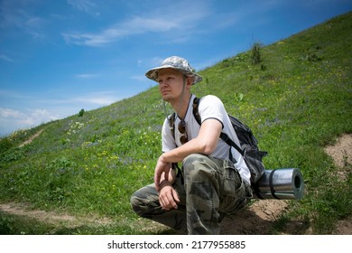White backpacker man wearing khaki panama and trousers sits on a hill looking for a direction to walk. Young hiker man in the mountains of Ile Alatau National Park, Kazakhstan. Male hiker on a walk. 