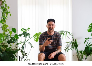 White Bald Young Woman With Hat Sitting Against A Window, Laughing While Messaging With His Phone.