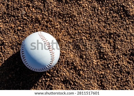A white baseball with red stitching sits on a textured dirt infield, bathed in sunlight