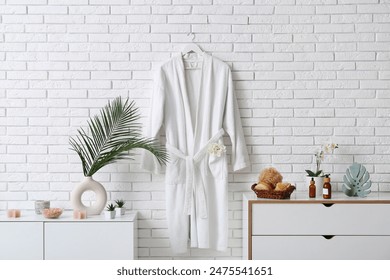 White bathrobe hanging on wall and different bath supplies on chest of drawers in bathroom