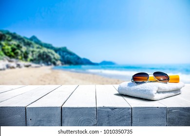 White Beach Towel With Sunglasses On White Wooden Table. Free Space For Your Decoration Of Product Or Text. Blurred Landscape Of Sea And Green Trees With Blue Sky.  