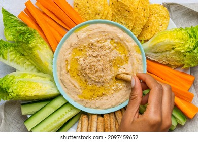 White Bean Hummus Alongside Cut Vegetables  A Woman's Hand Dipping The Vegetable Into The Bowl  Overhead View