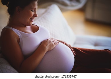 A white beautiful young pregnant woman sitting on a sofa and touching her baby bump with a happy smile of a future mother 