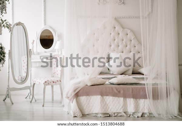 White bedroom interior with nobody. large cozy bed\
with a white canopy and oval dressing mirror with dressing table\
next to it