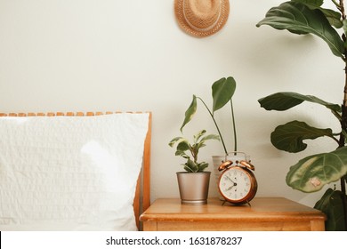 White Bedroom With Orange Wooden Bed, White Bedding, Night Stand And House Plants Near The Bed, Straw Hat On The Wall And Vintage Copper Alarm Clock On Night Stand