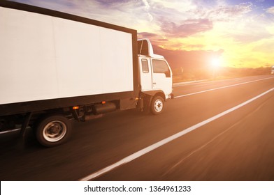 A white big box truck with space for text driving fast on the countryside road with trees and bushes against a night sky with sunset
