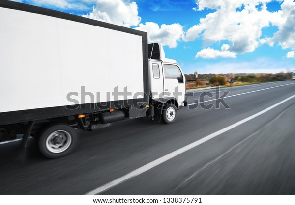 A white big box truck with space for text driving\
fast on the countryside road with trees and bushes against a blue\
sky with clouds