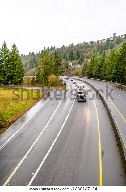 White big rig heavy-duty long haul diesel semi truck\
with spoiler transporting commercial cargo in covered semi trailer\
running on the turning divided wet slippery road in Columbia Gorge\
after rain