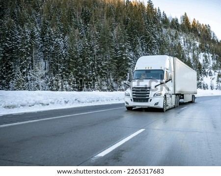 White big rig industrial semi truck transporting cargo in dry van semi trailer running on the winding winter dangerous slippery road with snow and ice and with snowy trees on the hill in Montana