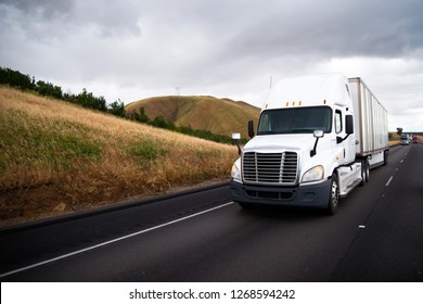 White Big Rig Semi Truck With Dry Van Semi Trailer Driving In Front Of Another Traffic Of Semi Trucks On The Straight Interstate Highway With Yellow Hill Roadside In California With Stormy Clouds