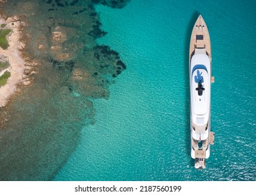 White big yacht for a billionaire anchored off a rocky shore aerial view. Mega yacht on blue water aerial view. Modern big white yacht on transparent water, coastline top view.