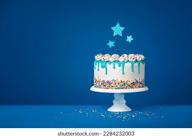White birthday drip cake with teal ganache, star toppers over dark blue background