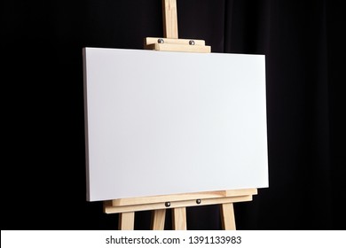 White Blank Canvas Stands On A Wooden Artistic Easel On Black Curtain Background. Horizontal Rectangular Mockup Canvas Wrapped On Stretcher Bar