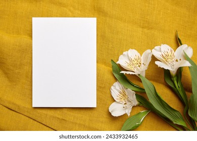 A white blank card ready for a personalized message, flanked by delicate white Alstroemeria flowers with speckled petals, resting on a textured mustard-yellow fabric backdrop - Powered by Shutterstock