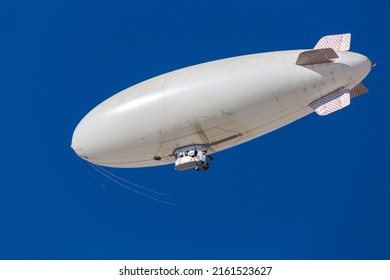 A white blimp, airship, or dirigible flying in blue sky. Close up detail of an unmarked zeppelin like flying vehicle. Floating high above in clear skies. Room for copy and or logo on side of airship.