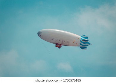 A White Blimp Flying In The Air With A Blue Sky Behind It.