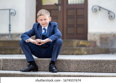 White Blond 9 Year Old Boy In A Blue Suit Sitting On The Stairs In Front Of A Church After His Communion