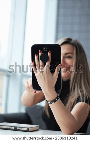 White blond girl holds a black tablet with the gadget holder (pop socket) and makes selfies. Woman is communicating online from a cafe or coworking space. Girl os on a video call.