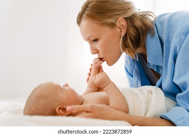 White Blonde Woman Kissing And Swaddling Her Baby In Bedroom At Home