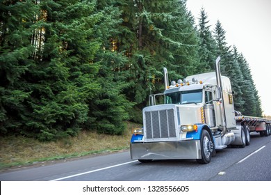 White and blue big rig classic American idol bonnet semi truck with vertical exhaust pipes transporting empty flat bed semi trailer driving on the road with frosted winter evergreen trees on the side