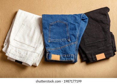 White blue black jeans pants stacked. Assortment of different color denim clothing in store shop. White denim jean pants, blue jeans, black denim. Top view on brown table