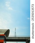 A white, blue sky, an orange Light Rapid Transit (LRT) station, and a bridge in the distance, with one black cell tower on top of it.