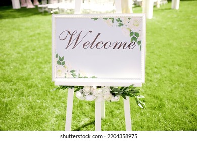 A White Board With The Inscription Welcome Stands On A Green Lawn. Signboard With The Invitation Of Guests To The Wedding Ceremony And Banquet. Beautiful Wedding Decor Decorated With Flowers