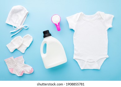 White Bodysuit, Hat, Mittens, Socks, Plastic Liquid Detergent Bottle And Pink Cup Or Scoop Of Powder On Light Blue Table Background. Pastel Color. Closeup. Preparing For Baby Clothes Washing.