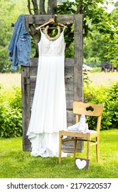 White boho wedding dress hanging on the tree in front of a door that is decorated with a blue jeans jacket; next to the wedding dress there are white sneakers standing on a wooden retro chair. - Shutterstock ID 2179220537