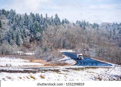 White bonnet popular professional big rig semi truck with bulk semi trailer going on the wet dangerous slippery icy winter road with snow on the trees and the sides of the winding highway