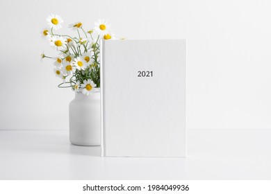 White book mockup, diary for 2021, white table, white chamomile flowers in vase. Front view. Place for text, copy space, mockup