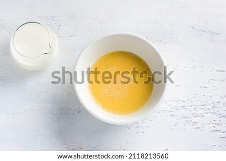 A white bowl with a beaten egg and a jug of milk on a light blue background, top view. Cooking omelet, homemade healthy food