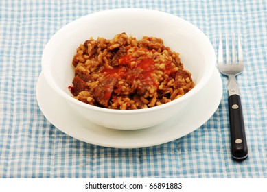 White Bowl Filled With A Cajun Cuisine Favorite --  Red Beans, Rice And Chunks Of Sausage Topped With Spicy Louisiana Hot Sauce.  All Setting On Blue Gingham Place Mat With Old Fashioned Fork.