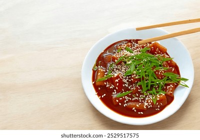 a white bowl filled with Tteokbokki and vibrant red sauce, garnished with sesame seeds and fresh herbs. Chopsticks rest on the bowl's edge, set on a light wooden surface, creating an appetizing scene. - Powered by Shutterstock