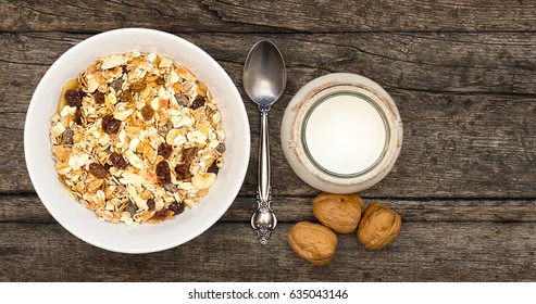 White Bowl With Granola Or Muesli And A Jar Of Milk Or Plain Yogurt With A Spoon On A Vintage Wood Background. Healthy Breakfast Top View Composition With Copy Space.
