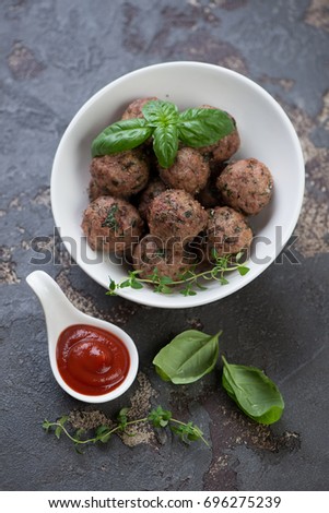 White bowl with oven-baked meatballs, fresh green basil, thyme and sauce. Vertical shot on a brown stone background