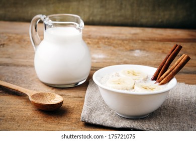 a white bowl of porridge with banana slices and cinnamon sticks, wooden spoon and a milk jug on rustic table  - Powered by Shutterstock