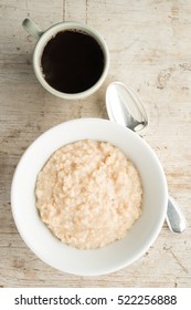 A White Bowl Of Porridge Oats Or Oatmeal On A Natural Wooden Background. Shot Overhead With Black Coffee And Silver Spoon