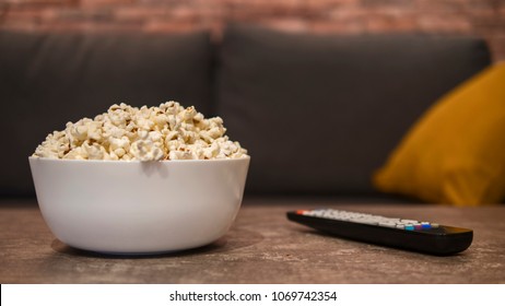 White bowl of white salty popcorn and remote control on a wooden table in front of a couch. Front view, blurry background