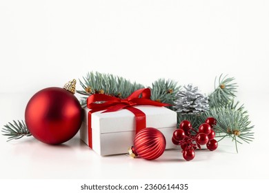 A white box with a gift tied with a red ribbon, a spruce branch and obligatory decorations on a white background.