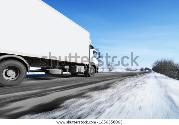 White box truck driving fast on the\
countryside winter road with snow against blue\
sky
