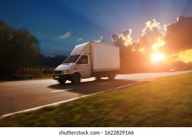 A White Boxed Truck Driving Fast On The Countryside Road With Green Trees And Bushes Against A Night Sky With Sunset