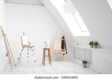 White and bright studio with a window and gray floor. Workspace of the artist. Easel, canvases and plaster figures for learning to draw. Conceptual interior without people.