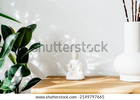 White Buddha statuette home decoration and clay vase standing on wooden cabinet. Minimalist design with green plants and white wall with shadows and highlights. Interior design decor. Selective focus.