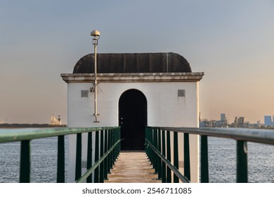 A white building with a domed roof stands at the end of a green-railed pier extending over the water. The evening sun casts a warm glow, while a cityscape is visible in the background across the water - Powered by Shutterstock