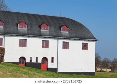 A white building featuring multiple red framed windows and doors with a distinctive black curved roof, set against a clear blue sky and green grass.