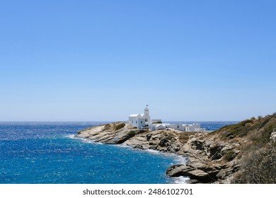 A white building sits atop a rocky coastline, surrounded by clear blue waters and under a bright blue sky. The famous Chrisopigi Monastery in Faros, Sifnos Greece - Powered by Shutterstock